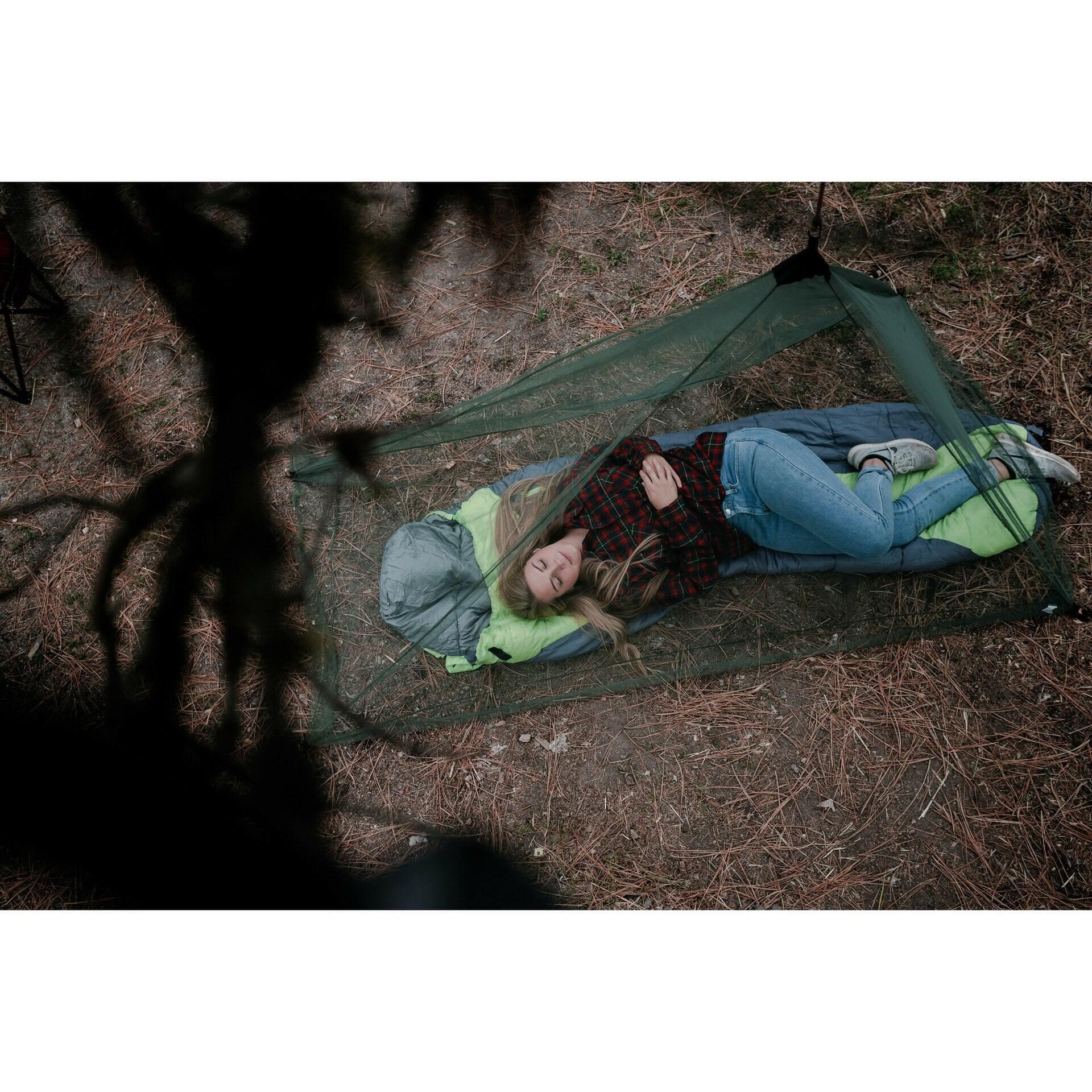 Woman sleeping in sleeping bag under green mosquito net outdoors on forest ground