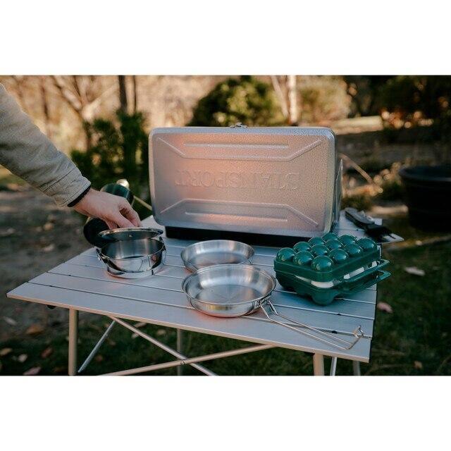 Outdoor camping table with stove, metal cookware, and green egg carrier case