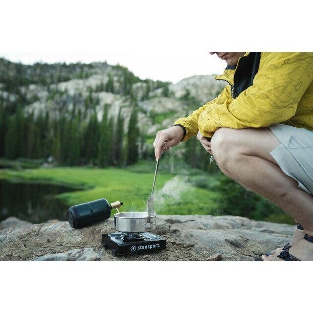 Person using Stansport stainless steel cooking utensils on a camping stove outdoors by a lake