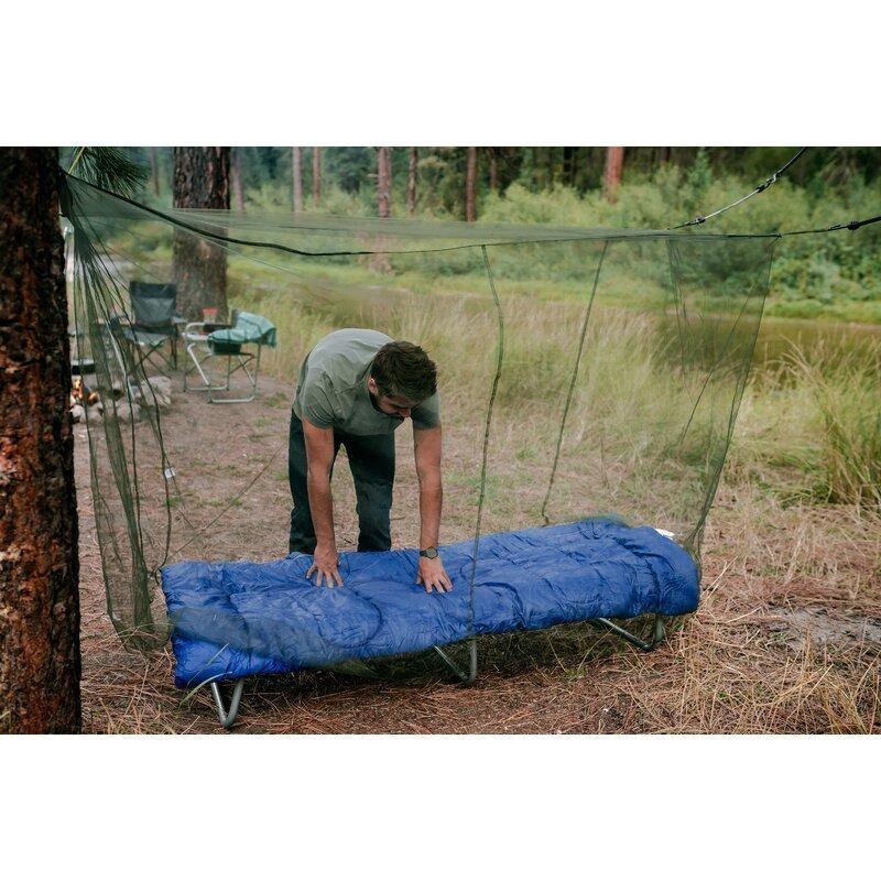 Man setting up suspension mosquito net over blue camping cot outdoors by river