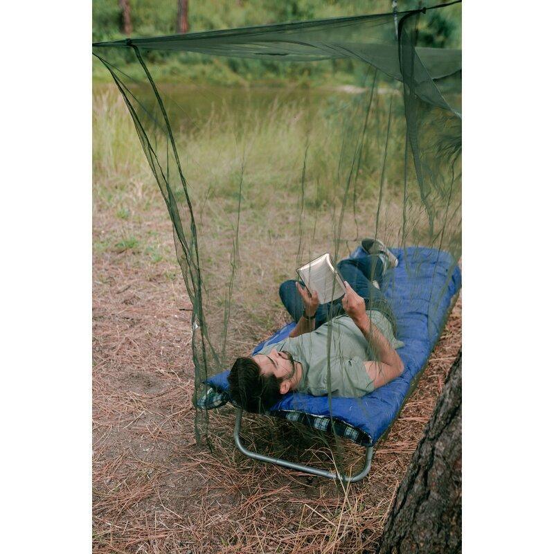 Man reading on cot under suspension mosquito netting outdoors in forest campsite
