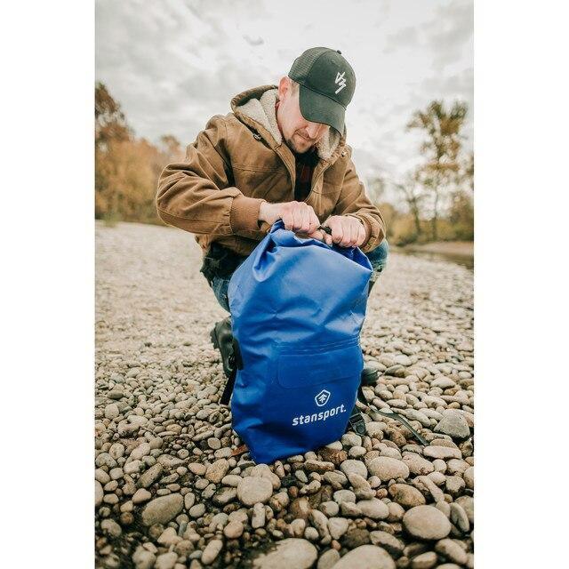 Man outdoors kneeling on rocky riverbank with blue Stansport waterproof dry bag