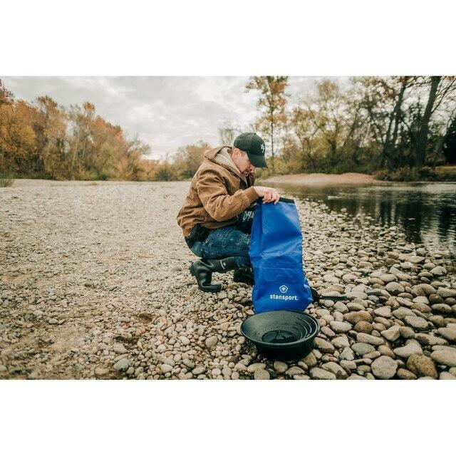Man outdoors by a river using Stansport blue waterproof dry bag on rocky shoreline