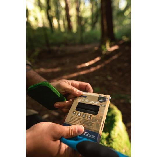 Hands holding Stansport solid fuel sticks box in a forest setting, outdoor camping gear