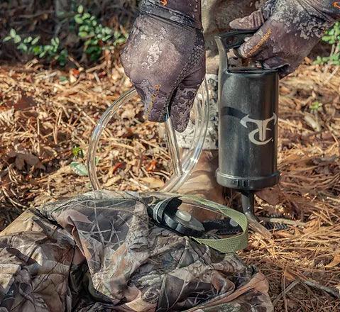 Hands in camo gloves inflating compact camouflage ground blind outdoors with pump