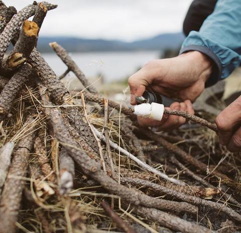 Hands using Zippo emergency fire kit to light kindling sticks outdoors by water